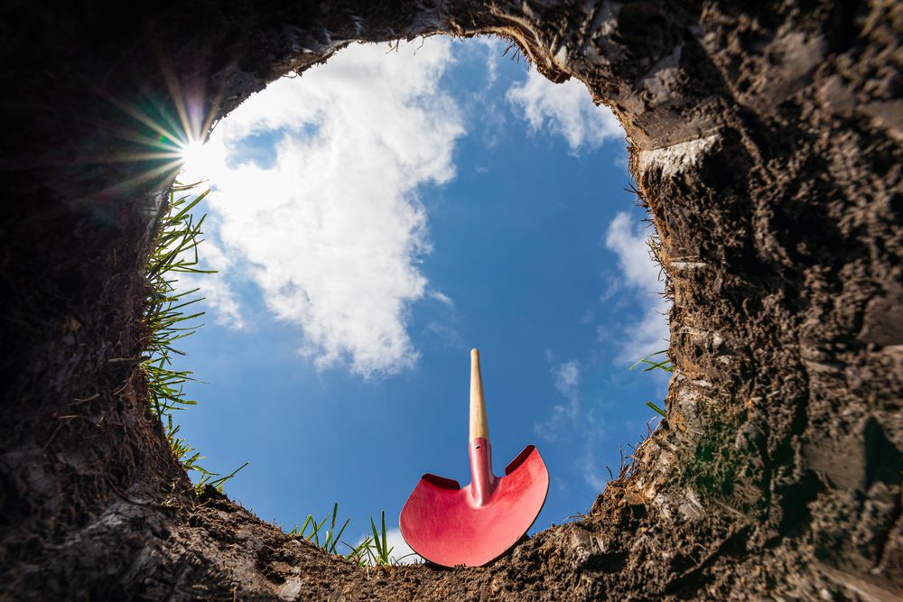 Et stort hul i jorden - set nedefra og op mod den blå himmel. En rød spade ses i kanten af hullet
