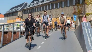 Folk cykler på en solrig dag ad en afmærket cykelsti i et byområde med en murstensbygning i baggrunden og andre cyklister og fodgængere i nærheden.