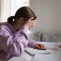 Teenager fotograferet fra siden sidder ved et skrivebord og læser lektier med hovedet i hænderne. Hun har det mørke hår sat op i en hestehale og har en violet hættetrøje på.