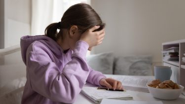 Teenager fotograferet fra siden sidder ved et skrivebord og læser lektier med hovedet i hænderne. Hun har det mørke hår sat op i en hestehale og har en violet hættetrøje på.