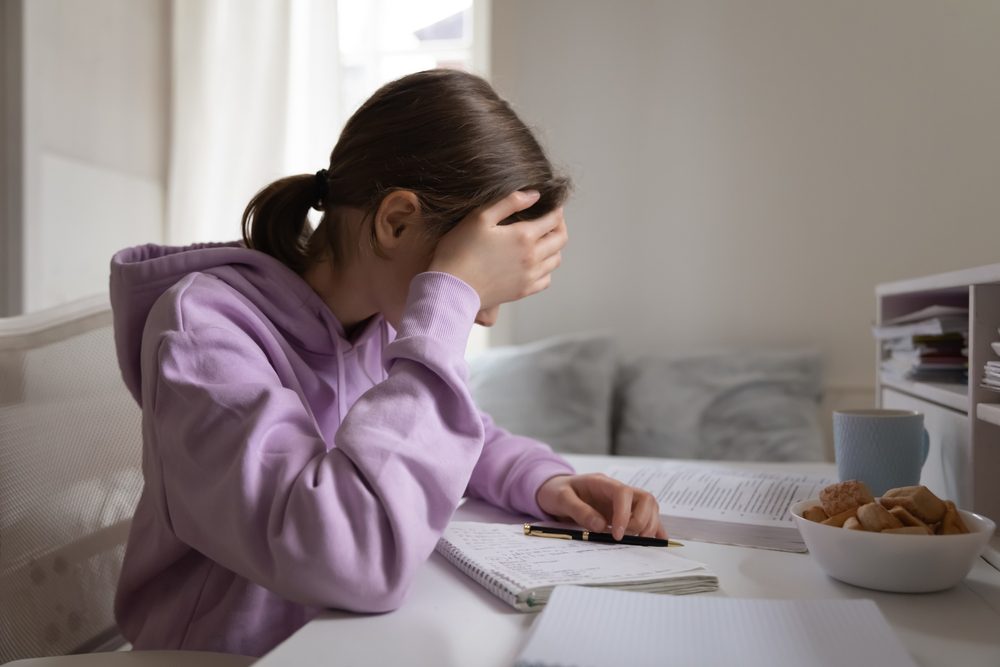 Teenager fotograferet fra siden sidder ved et skrivebord og læser lektier med hovedet i hænderne. Hun har det mørke hår sat op i en hestehale og har en violet hættetrøje på.
