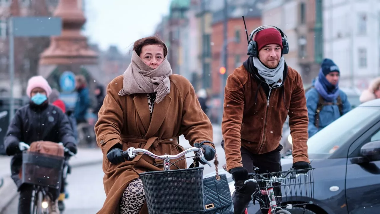 En kvinde og en mand pakket ind i halstørklæder og frakker cykler på en gade i byen i koldt vejr. Andre cyklister og fodgængere er synlige i baggrunden, nogle iført masker og vintertøj.