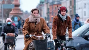 En kvinde og en mand pakket ind i halstørklæder og frakker cykler på en gade i byen i koldt vejr. Andre cyklister og fodgængere er synlige i baggrunden, nogle iført masker og vintertøj.
