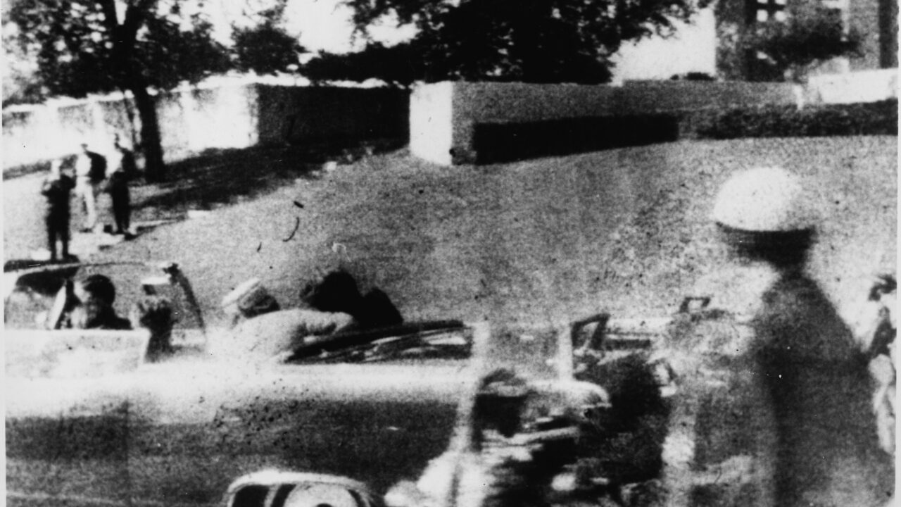 Limousinen med blandt andre USA's præsident John F. Kennedy i sekunderne efter, at et dødbringende skud har ramt ham i hovedet. Omkring 50 vidner forklarede bagefter, at de havde hørt skud eller set aktivitet på græshøjen bagest i billedet. (Foto: Mary Moorman)
