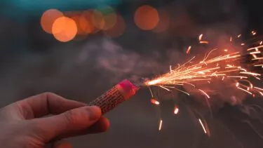 En hånd, der holder en tændt stjernekaster, med klare gnister, der flyver, og varme, slørede orange lys i baggrunden, der skaber en festlig og fejrende atmosfære.