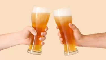 To hænder skænker høje ølglas mod en almindelig beige baggrund. Hvert glas er fyldt med gyldent øl og en skummende skumkrone.