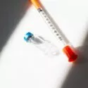 En sprøjte med orange hætte ved siden af et lille glasglas med blåt låg, begge placeret på en hvid flade med skygger, der delvist dækker dem.