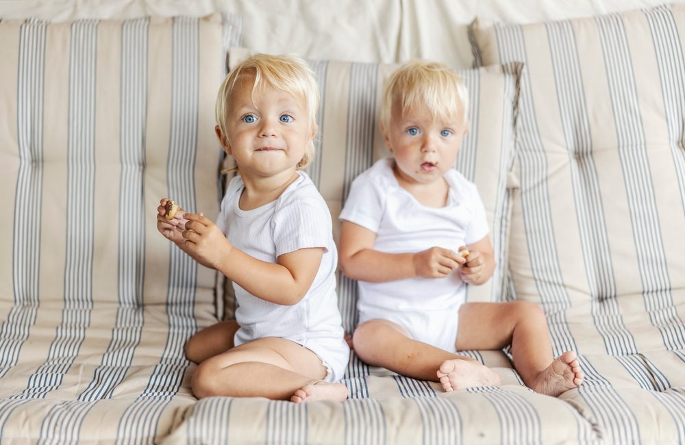 To hvidblonde rollinger på omkring et år sidder i en sofa og kigger på kameraet