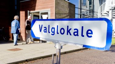 Et skilt med ordet Valglokale peger mod en indgang til en bygning, hvor to personer står ved døren, hvilket indikerer et valgsted i Norge.