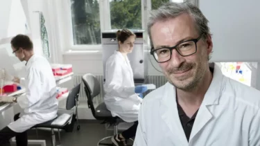 Tre forskere i hvide kitler arbejder i et lyst laboratorium. To sidder ved arbejdsstationer med laboratorieudstyr, mens en mand med briller står i forgrunden og smiler til kameraet.