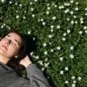 En person med lukkede øjne ligger på grønt græs omgivet af små hvide blomster og nyder sollyset med hænderne hvilende bag hovedet. Scenen formidler afslapning og ro i naturen.