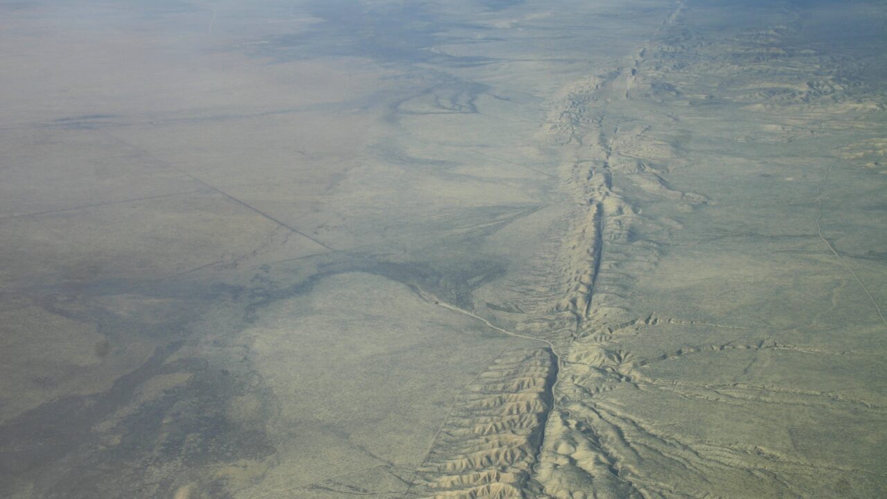 Luftfoto af et tørt, fladt landskab med en lang, takket forkastningslinje, der skærer gennem terrænet og viser højderygge og sprækker i den tørre jord. Sparsom vegetation og subtile farvevariationer er synlige.