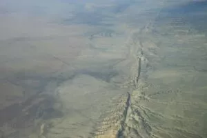 Luftfoto af et tørt, fladt landskab med en lang, takket forkastningslinje, der skærer gennem terrænet og viser højderygge og sprækker i den tørre jord. Sparsom vegetation og subtile farvevariationer er synlige.