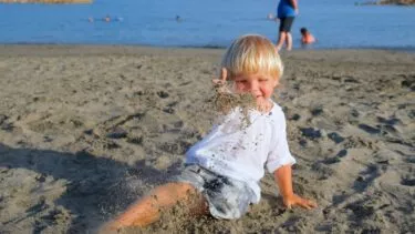 Et lille barn med lyst hår, iført hvid skjorte og shorts, sidder på en sandstrand og kaster legende med sand. Havet og nogle få mennesker er synlige i baggrunden under en klar himmel.