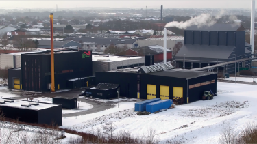 Et moderne industrianlæg, Thisted geotermin-anlægget, med sorte bygninger og gule accenter ligger i et snedækket landskab. Røg stiger op fra en høj skorsten, og en by med mange huse er synlig i baggrunden.