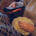 Closeup af hænder, der spiser popcorn og holder på en fjernbetjening samtidig.