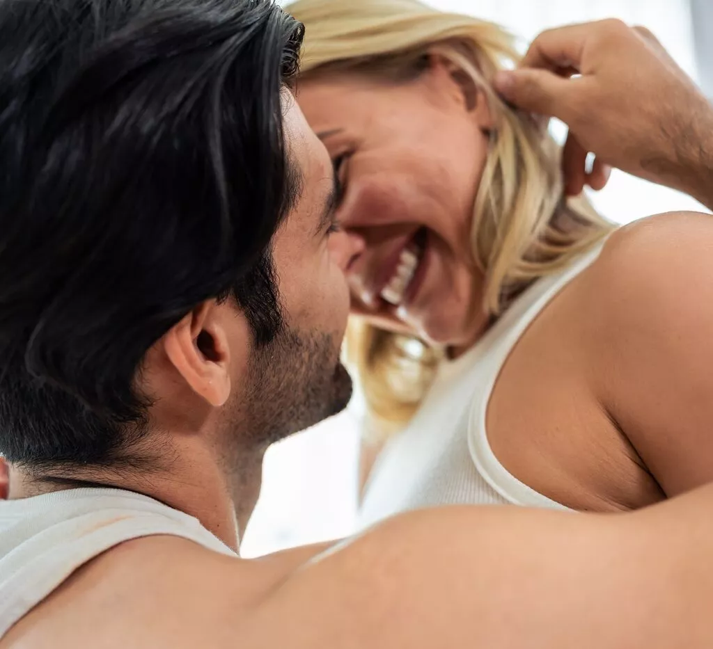 Et nærbillede af et smilende par, der omfavner hinanden intimt og deler et legende øjeblik, hvor manden forsigtigt rører ved kvindens ansigt, mens hun griner - begge iført hvide toppe i et lyst, indendørs miljø, der udstråler sexappeal.