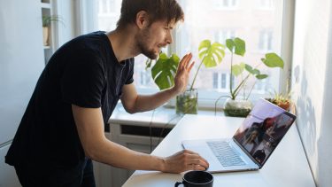 Mand står ved sin laptop og vinker akavet til deltagerne i et videomøde