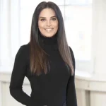 En kvinde med langt, glat, brunt hår iført sort rullekrave står indendørs foran store, lyse vinduer og smiler til kameraet.