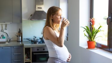 Lyshåret ung kvinde står i et køkken og drikker et glas postevand
