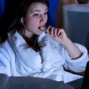 Træt kvinde iført badekåbe spiser snack og drikker rødvin sent om aftenen foran computeren. (Foto: Shutterstock)