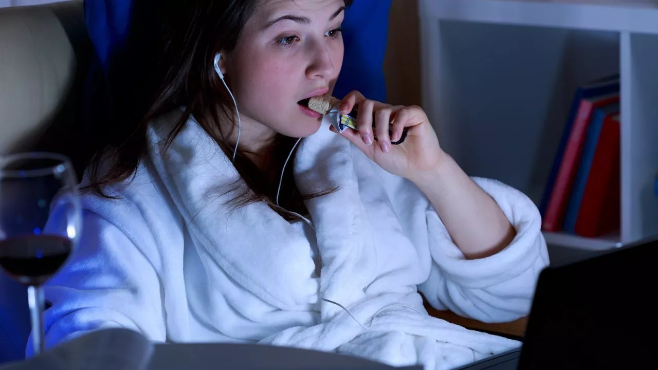 Træt kvinde iført badekåbe spiser snack og drikker rødvin sent om aftenen foran computeren. (Foto: Shutterstock)
