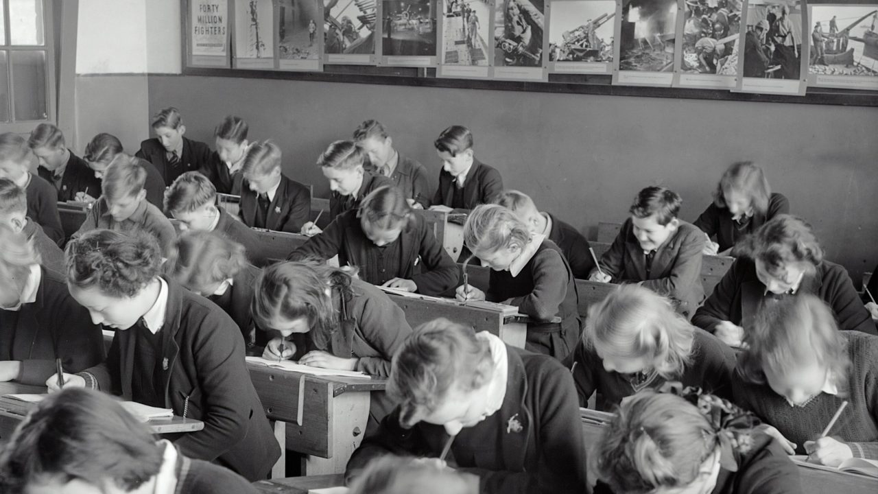 Sort/hvid-billede af skoleelever, der sidder med hovedet ned i bordet, tilsyneladende meget optagede af deres skoleopgaver.