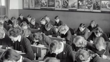 Sort/hvid-billede af skoleelever, der sidder med hovedet ned i bordet, tilsyneladende meget optagede af deres skoleopgaver.