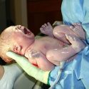 Nyfødt, skrigende baby, der stadig er dækket i fosterfedt, som bliver holdt af en person med plastikhandsker og blå papirkittel