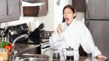 Kvinde, der står i et køkken, iført en hvid morgenkåbe og står med en kop kaffe i den ene hånd, mens hun gaber.