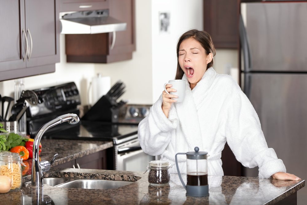 Kvinde, der står i et køkken, iført en hvid morgenkåbe og står med en kop kaffe i den ene hånd, mens hun gaber.