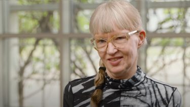 Portrætbillede af Lotte Bjerre Knudsen. Hun har gråligt hår i en fletning og pandehår, store briller og en højhalset sort og hvid bluse på.