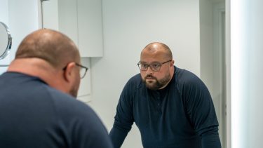 Midaldrende mand med nakkedelle og måne, briller og fuldskæg står lænet ind over vasken og kigger sig i spejlet. Han ser småbekymret ud.