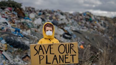 Barn i gul regnjakke foran affaldsbunke, der står med et skilt med ordene "save our planet"