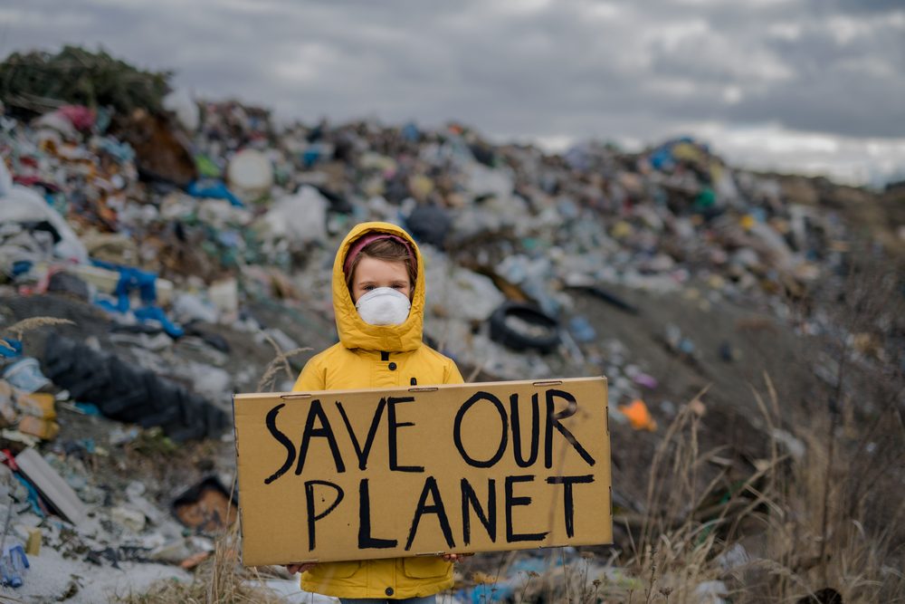 Barn i gul regnjakke foran affaldsbunke, der står med et skilt med ordene "save our planet"