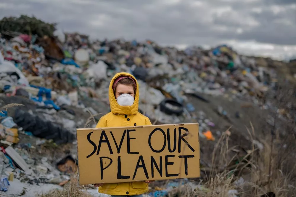 Barn i gul regnjakke foran affaldsbunke, der står med et skilt med ordene "save our planet"