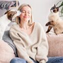 Kvinde i sofa mellem en hund og en kat, hunden slikker hende på kinden, katten vil også have opmærksomhed
