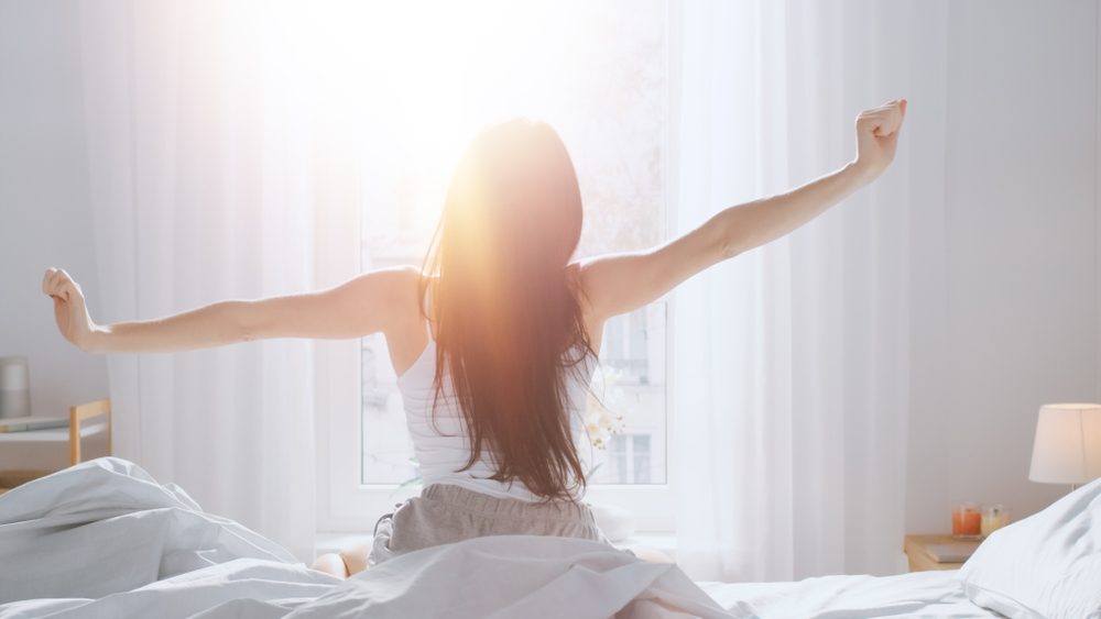 Mørkhåret kvinde, der trækker gardin fra om morgenen, hvor solen stråler ind