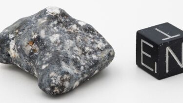 Til venstre ses en aubrit meteorit i en grå granitlignende farve ved siden af en terning