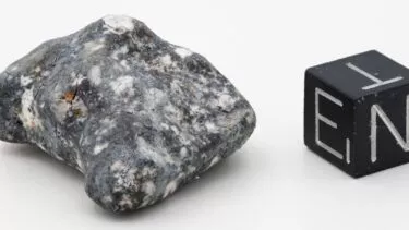 Til venstre ses en aubrit meteorit i en grå granitlignende farve ved siden af en terning