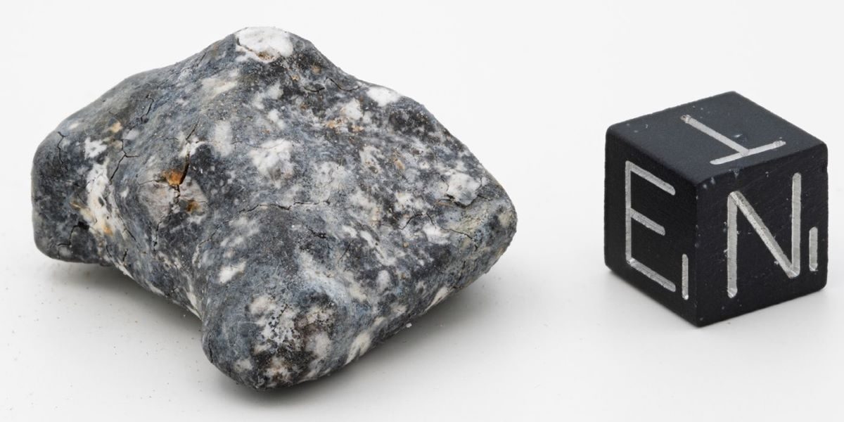 Til venstre ses en aubrit meteorit i en grå granitlignende farve ved siden af en terning