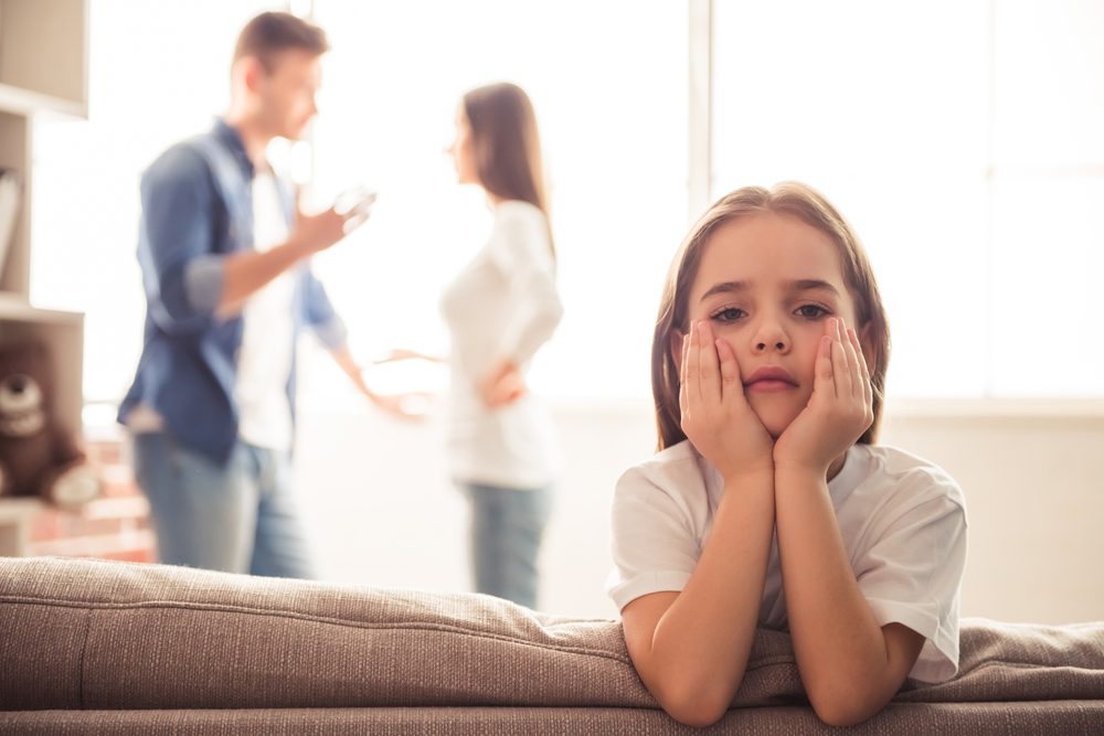 Pige sidder på kanten af sofa, mens far og mor skændes i baggrunden.