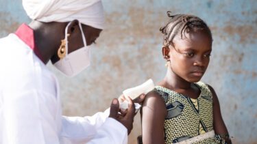 En ung sygeplejerske i en hvid frakke iført en maske sprøjter en intramuskulær vaccine ind i armen på en skolepige i en landsby i Afrika