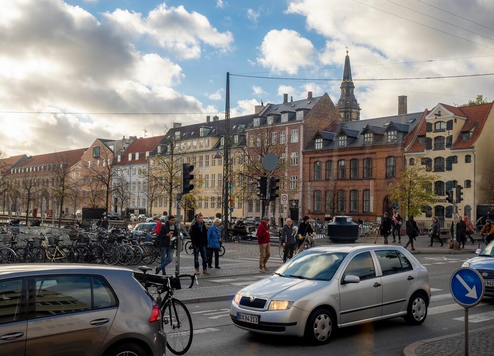 Biler kører ved Christianshavn i København, mens fodgængere venter ved en fodgængerovergang.