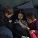 Tre små børn sidder i en sofa og kigger på den samme tablet.