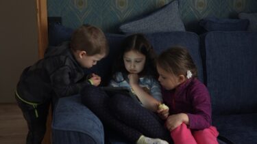 Tre små børn sidder i en sofa og kigger på den samme tablet.