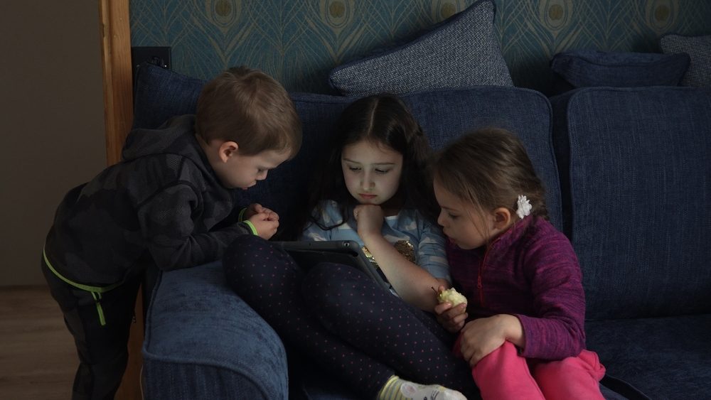 Tre små børn sidder i en sofa og kigger på den samme tablet.