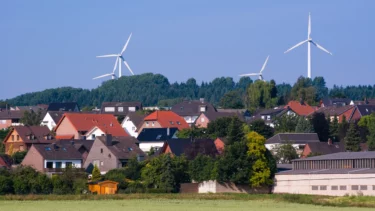 Tre vindmøller ses i horisonten bagved skov og huse