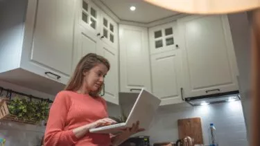 En kvinde står i sit køkken med sin computer