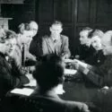 En gruppe mænd sidder rundt om et bord og kigger på en person, der sidder med ryggen til